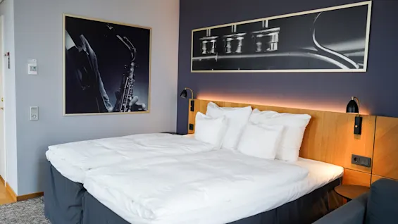 En pent dobbeltseng med hvitt sengetøy flankeres av to innrammede svart-hvitt-fotografier av musikkinstrumenter, plassert i et stilfullt, moderne rom med dempet belysning.