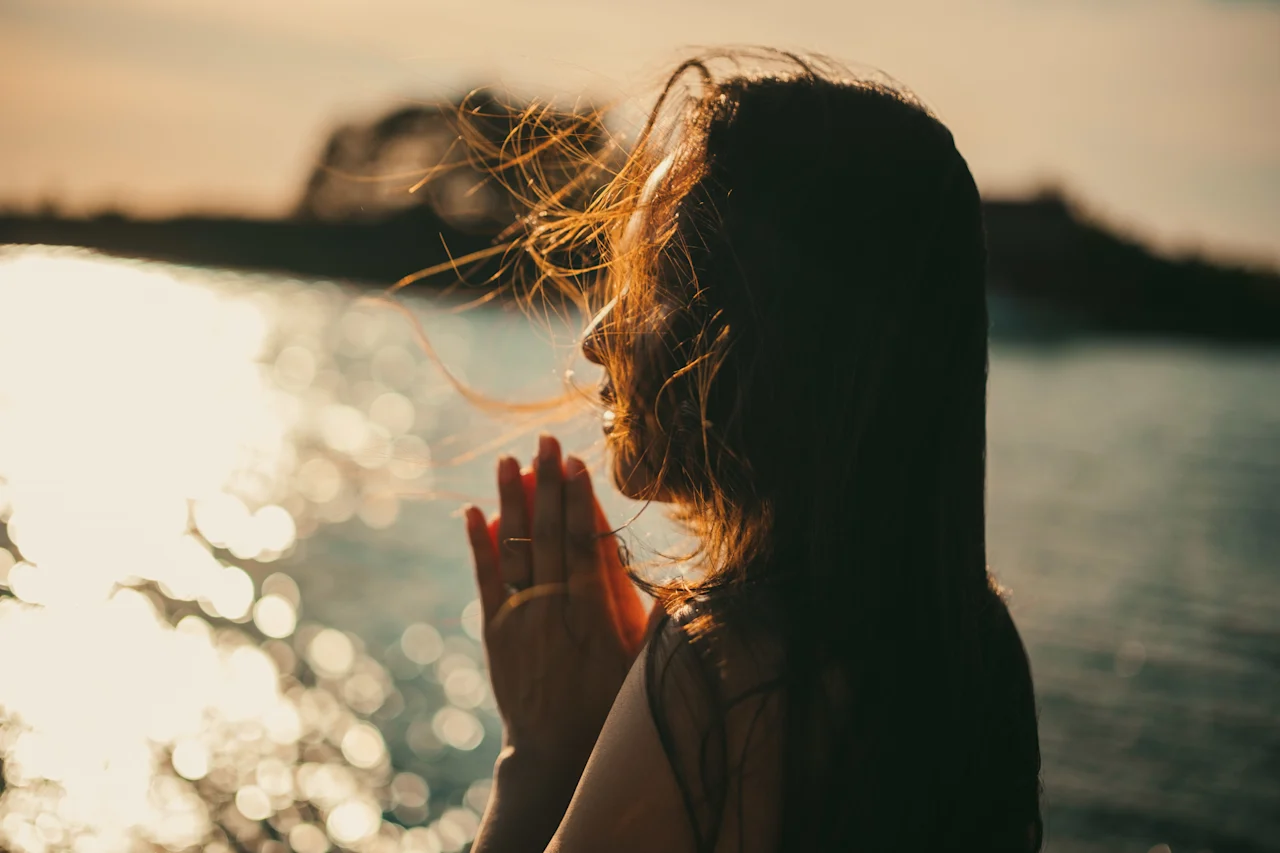 En person med hænderne knyttet foran sig, silhuet mod et glitrende vandområde ved solnedgang, med håret der blæser blidt.