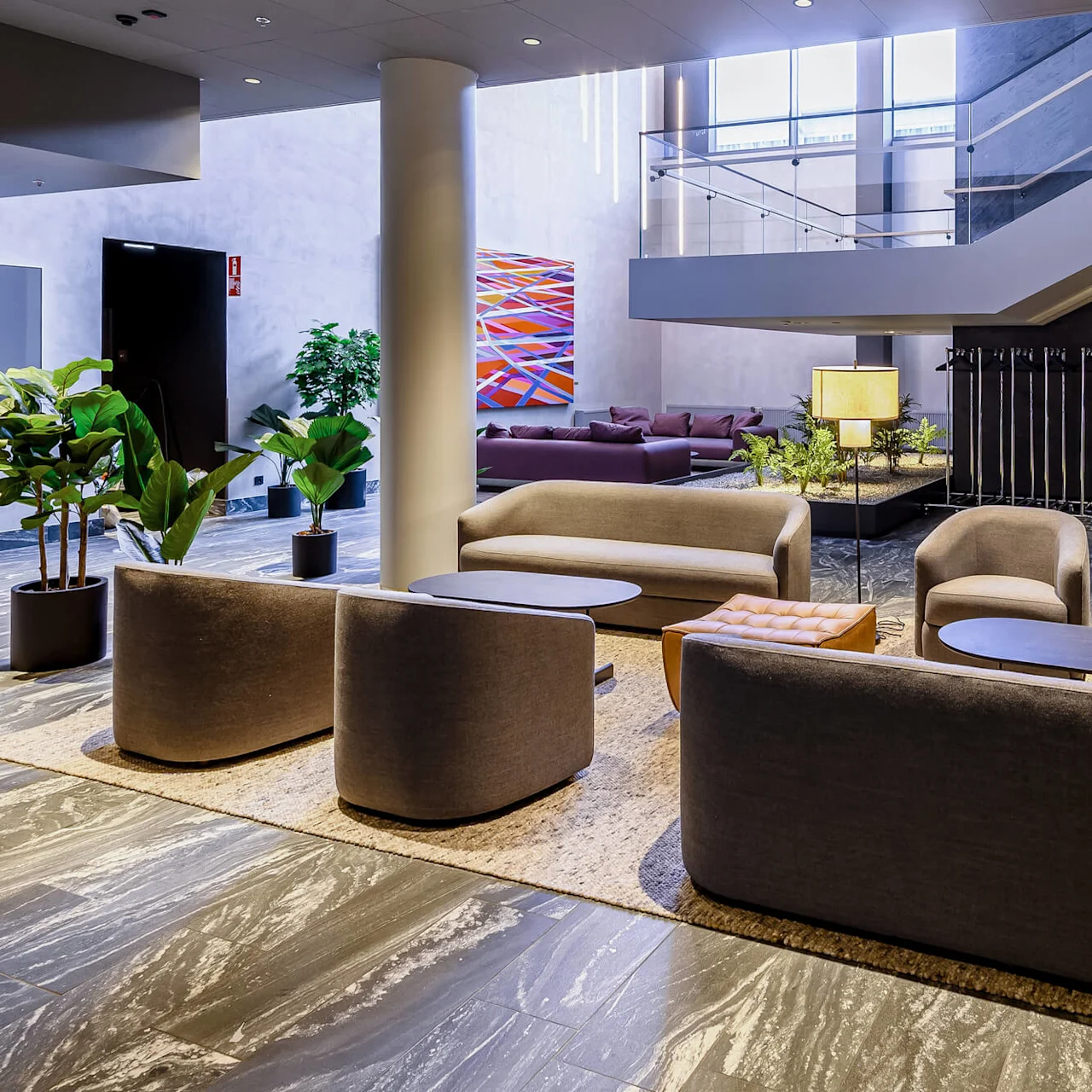 Seating area with gray chairs and tables arranged on a rug. Lamps and plants provide decor. A staircase and abstract painting are visible in the modern lobby.