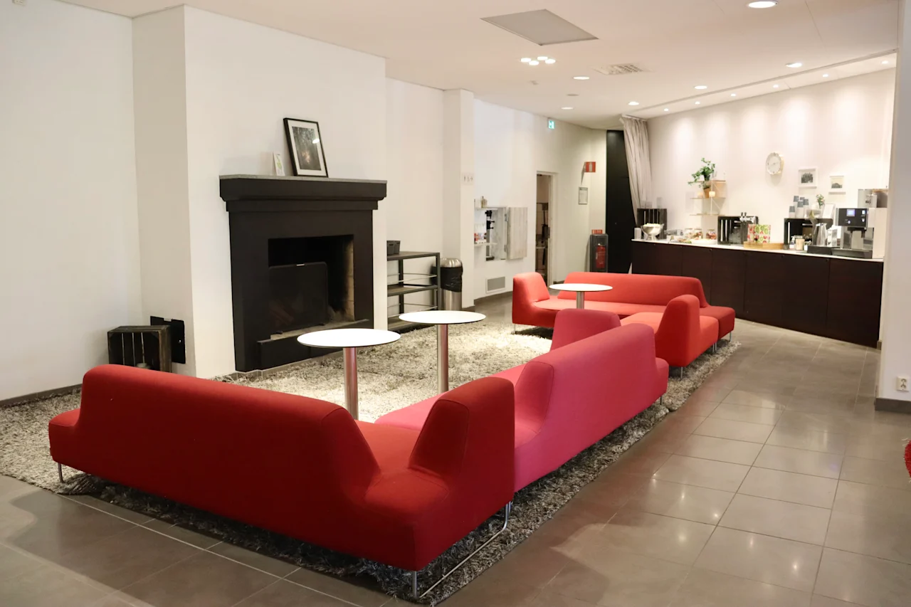 Red sofas face small round tables, arranged on a carpet in a lounge with a fireplace. Adjacent is a counter with coffee machines and cups against a tiled floor.