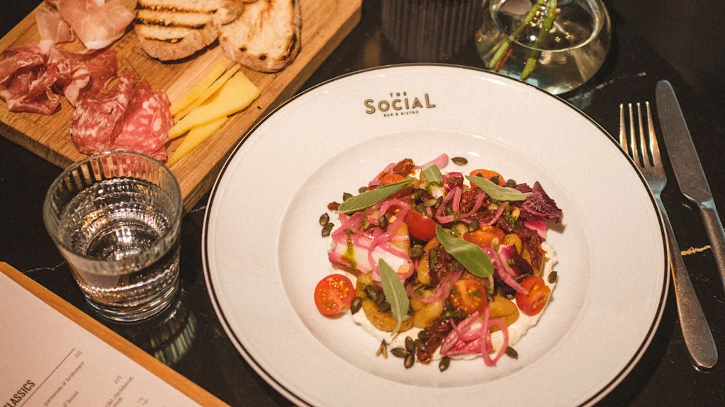 Et lækkert måltid serveret på et bord, med en farverig salat, charcuteri, ost, brød og en menu for THE SOCIAL BAR AND BISTRO.