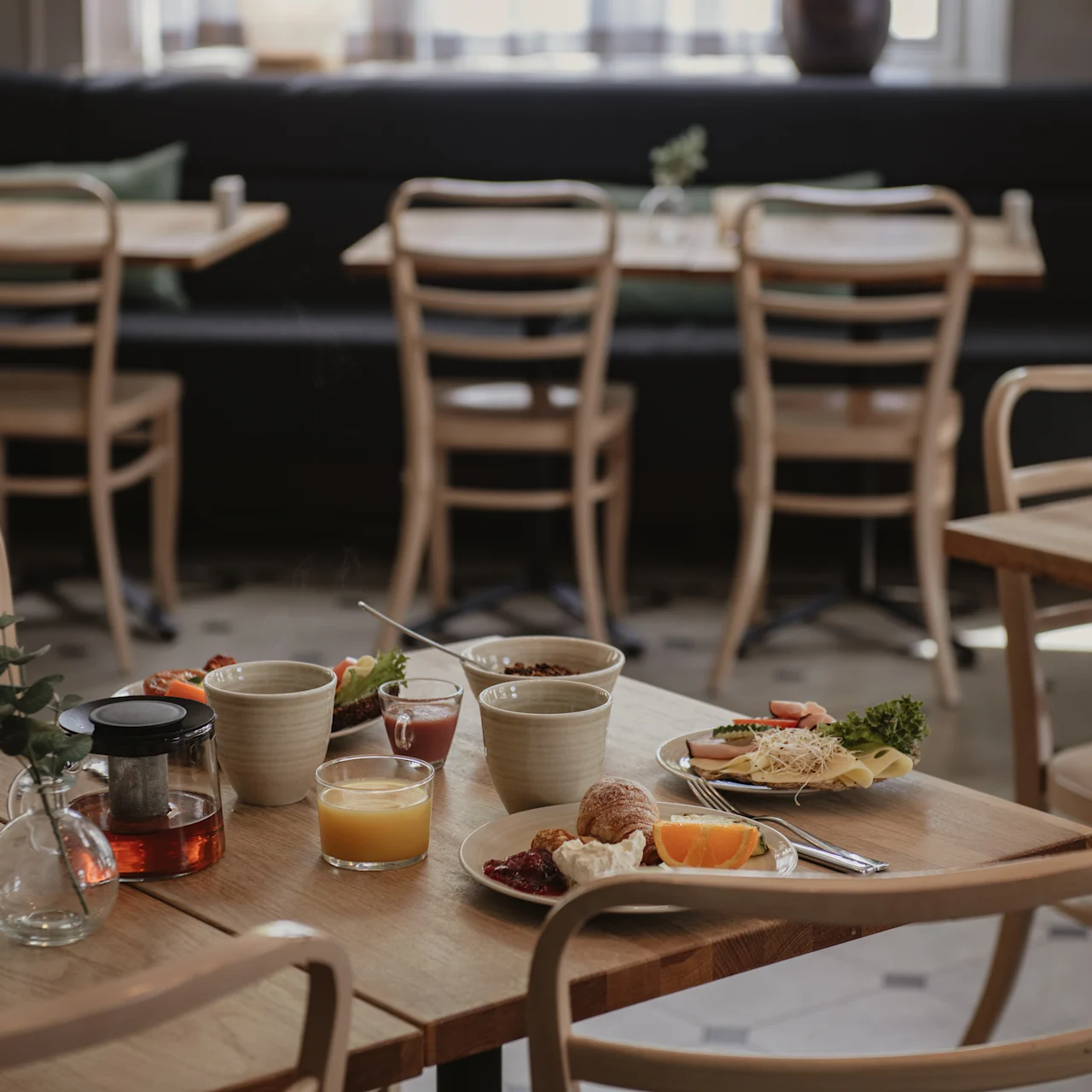 Ett bord har frukostprodukter, inklusive juice, te, bröd och frukt, i en mysig kafémiljö med trästolar och andra bord i bakgrunden.