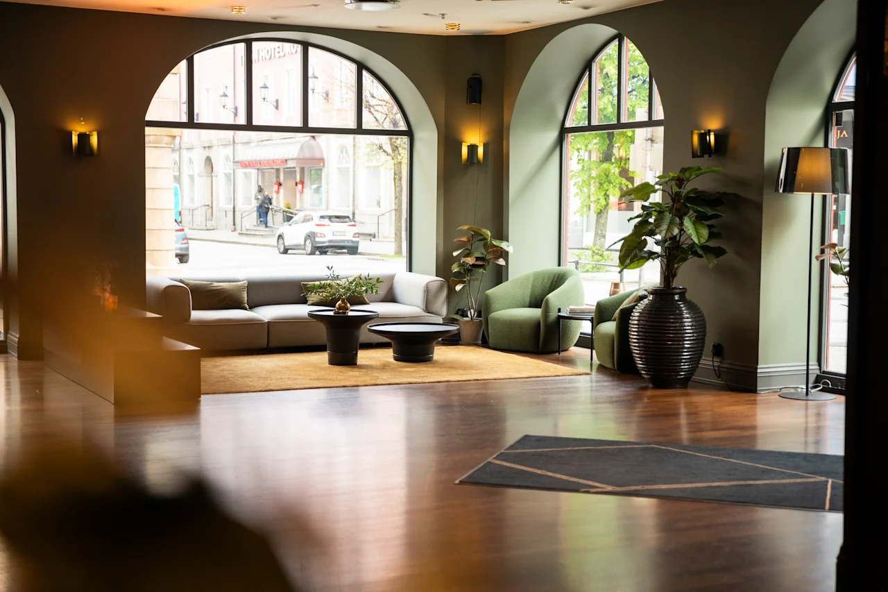 Et sitteområde med en moderne grå sofa og en grønn lenestol står i et rom med store buede vinduer, som vender ut mot en gate. Potteplanter gir grønt til innredningen.
