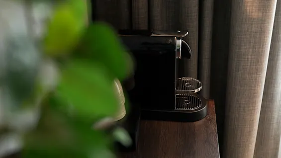 En elegant svart kaffemaskin på en mørk treoverflate, med grønt løvverk i forgrunnen og teksturerte gardiner bak.