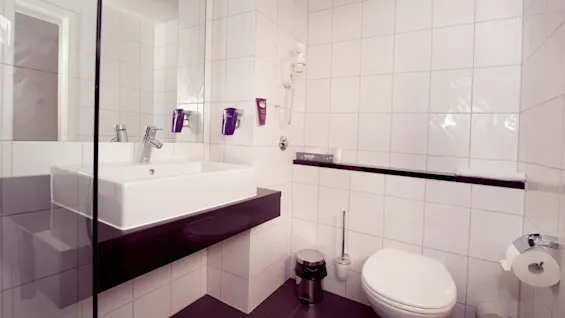 A modern bathroom with a white rectangular basin, wall-mounted faucet, shower, toilet, and accessories in a clean, tiled space.