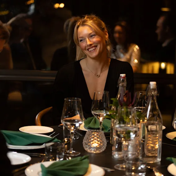 En person smiler lyst ved et middagsbord, og nyter et sosialt samvær på en restaurant.