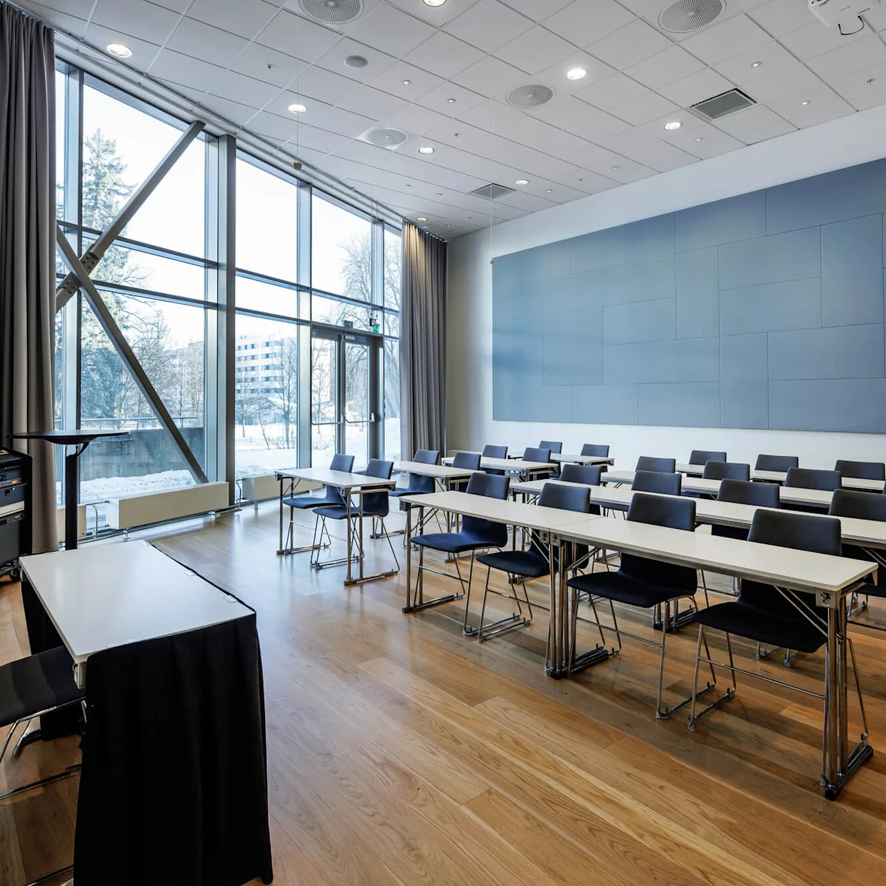 Classroom with rows of chairs and tables facing a large wall-mounted screen; wooden floors and large windows showing a snowy outdoor scene.