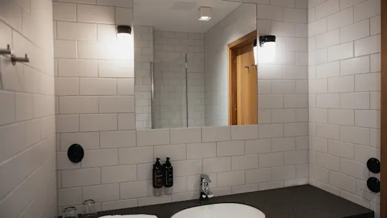 Moderni kylpyhuone, jossa valkoiset laatat, iso peili heijastaa puuovea, pesuallas ja hygieniatuotteita tummalla työtasolla.