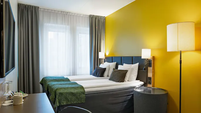 A neatly made double bed with white sheets and dark cushions is set against a yellow wall, adjacent to a large window with sheer curtains and a floor lamp.