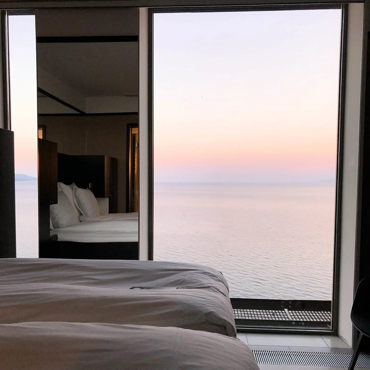 Senge er placeret i et moderne rum med et stort vindue, der afslører en fredfyldt udsigt over havet ved solnedgang. Bløde pastel farver oplyser himlen og vandet og skaber en beroligende atmosfære.
