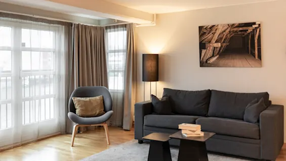 En koselig stue med en grå sofa, en lenestol og en gulvlampe, opplyst av naturlig lys fra store vinduer.