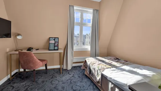 Ett fridfullt hotellrum med en enkelsäng, skrivbord och stol vid ett fönster, visar en ljus utsikt utomhus. Inga personer närvarande.