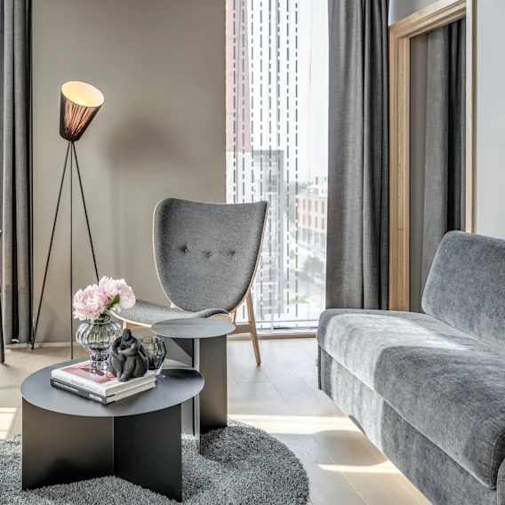 Grå sofa, rundt sofabord med blomster og lenestol er arrangert i en moderne stue. En gulvlampe står ved siden av stolen, nær store vinduer med byutsikt.