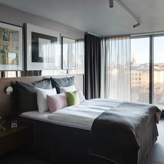 Ett modernt hotellrum med en bekväm säng, en stilfull fåtölj vid ett stort fönster och utsikt över staden.