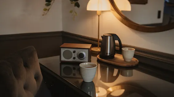 Viihtyisä hotellihuoneen työpöytä, jossa radio (FM AM, TUNING), vedenkeitin ja kaksi kuppia, valaistuna lämpimällä lampulla. Täydellinen rentoutumiseen.