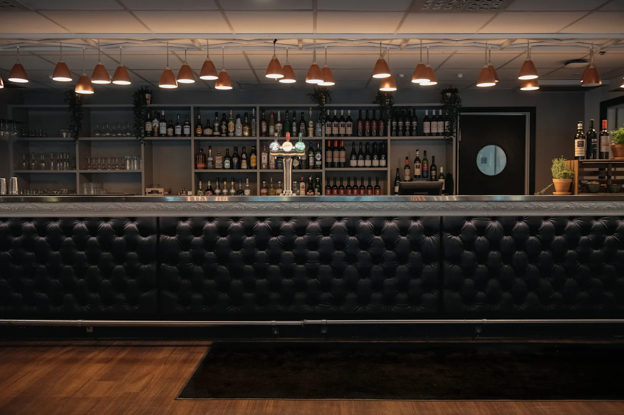 A stylish bar area with a black tufted counter, shelves stocked with bottles and glasses, and warm pendant lighting.