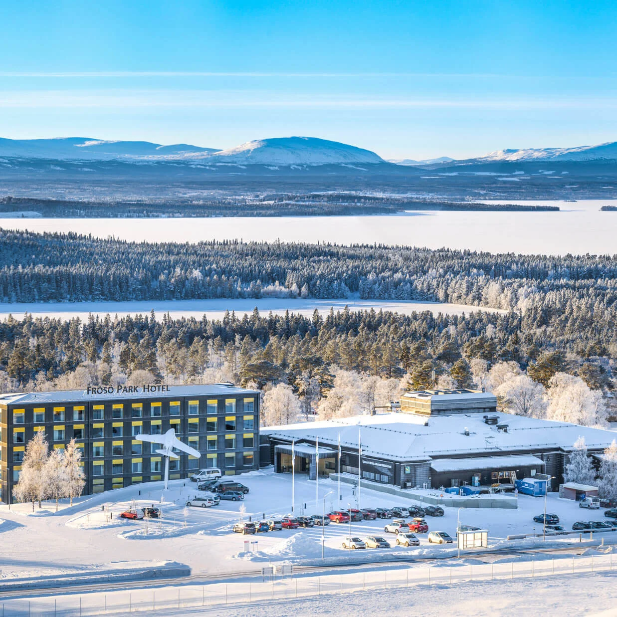 Frösö Park Hotel står täckt av snö, omgivet av frostiga träd och avlägsna berg under en klarblå himmel.
