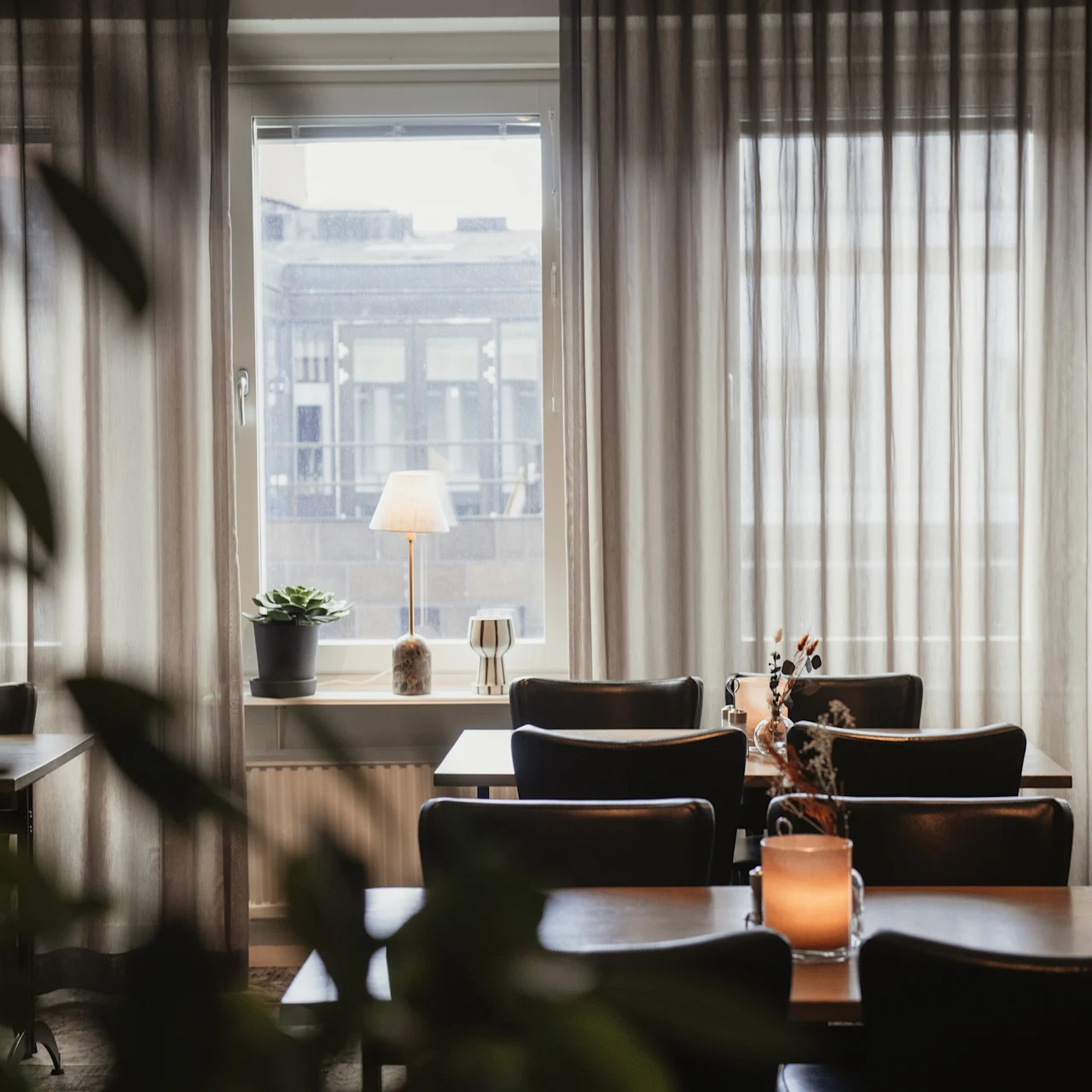 Dining tables are arranged with chairs, centered around a glowing candle in a softly lit restaurant. Large curtains cover windows, partially opened to city buildings outside.