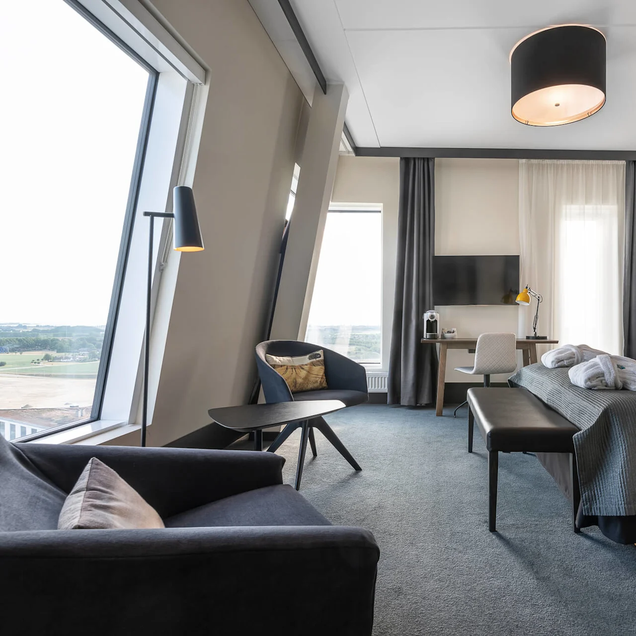 A modern hotel room features a large bed with pillows and a throw, a desk with a chair and lamp, and a seating area with armchairs near wide windows offering scenic views.