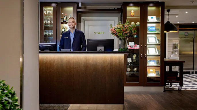 En smilende resepsjonist står ved en trepult, dekorert med blomster, i en hotellobby. Bak er en utstillingshylle med glass og priser. En dør merket "STAFF" er synlig.