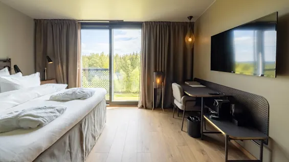 Ett modernt hotellrum med säng, skrivbord och platt-TV, med utsikt över träd genom ett stort fönster.