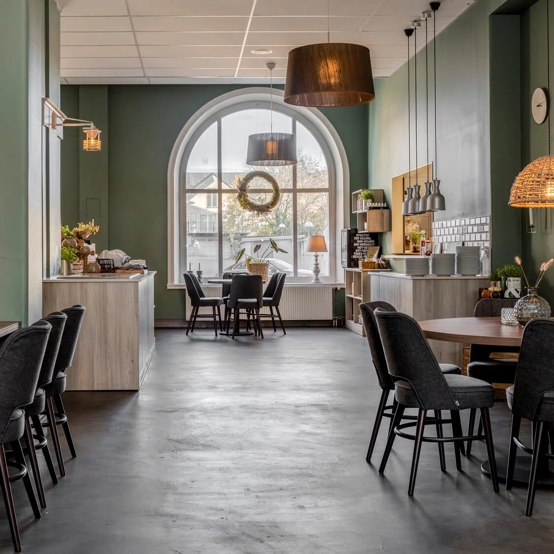 A cozy cafe interior with wooden tables, upholstered chairs, and decorative lighting, framed by large arched windows and green walls.