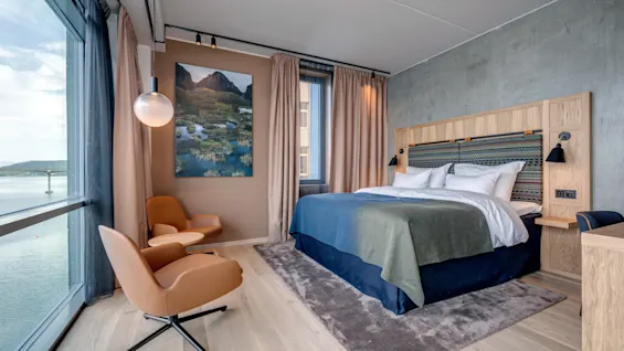 Et hyggeligt hotelværelse med en stor seng og hvidt sengetøj, to brune stole, et rundt bord og et landskabsmaleri. Rummet har udsigt over vand gennem et stort vindue.