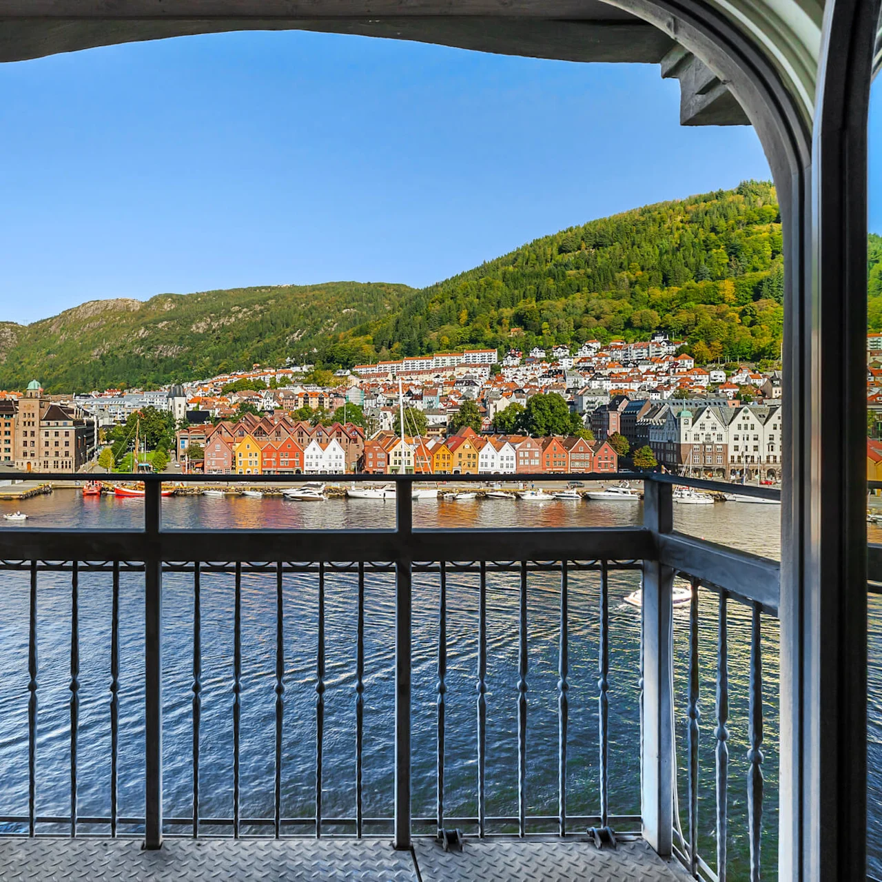 En balkong med utsikt over en havn med fargerike hus langs vannet. Fjell reiser seg i bakgrunnen under en klar blå himmel.