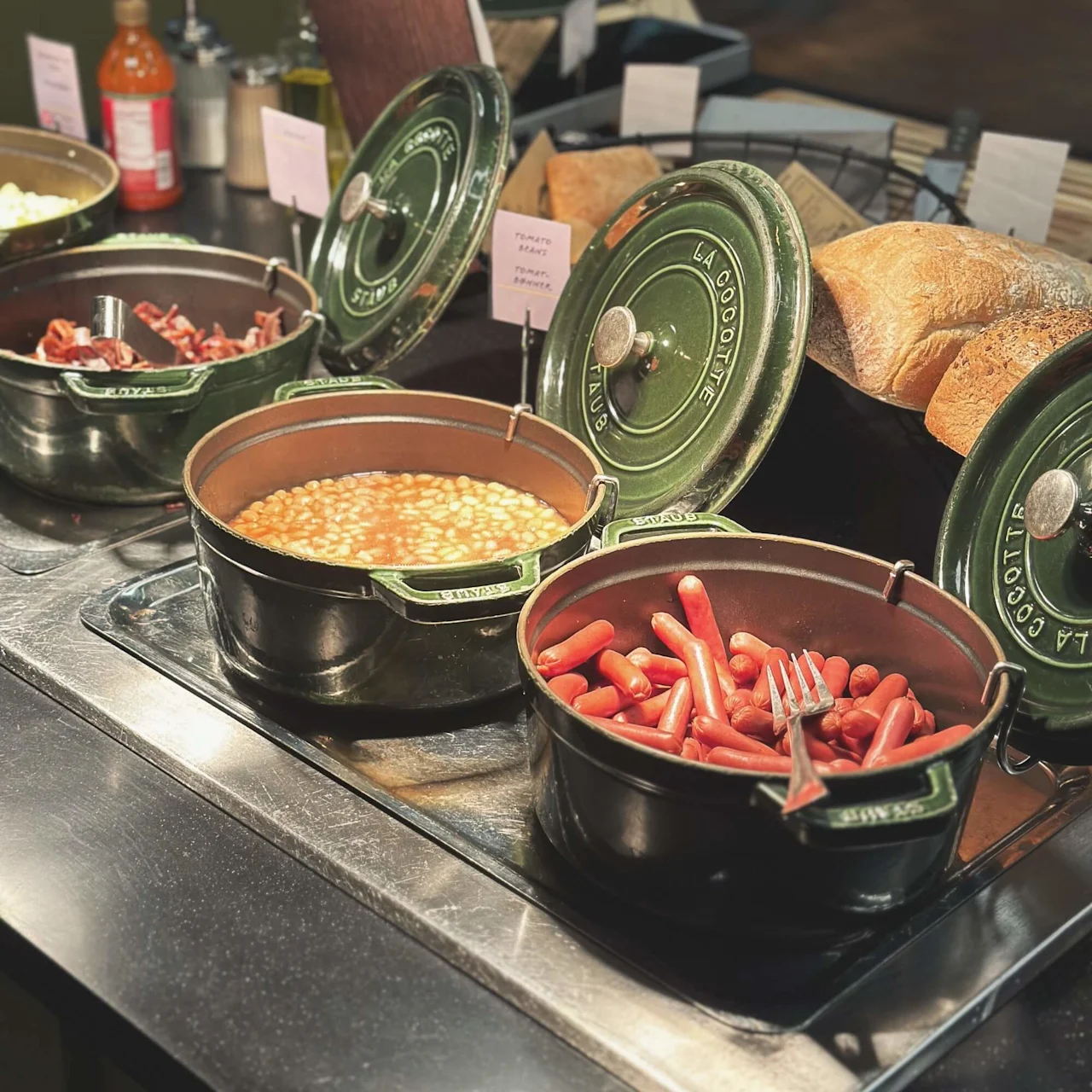 Large green pots filled with sausages and baked beans are displayed on a buffet table beside fresh bread. Signs read "Tomato harry tomats hanner." Bottles and jars are in the background.