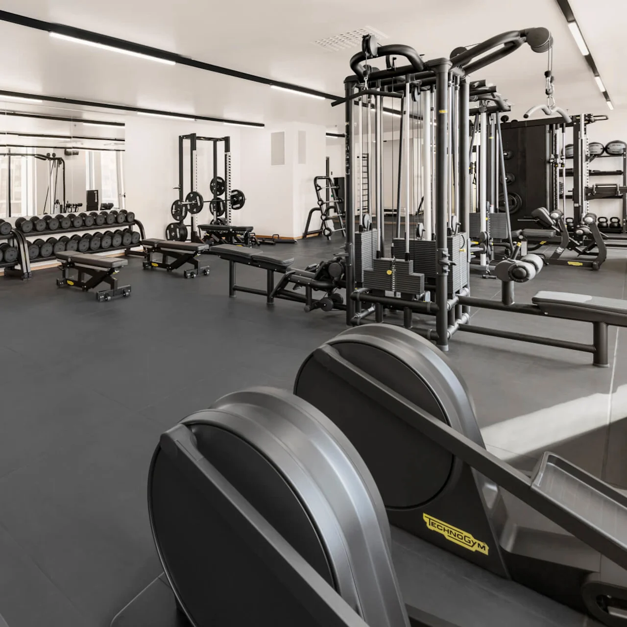Ett rymligt gym har olika styrketräningsmaskiner, bänkar, hantelställ och viktskivor, ordnade prydligt på ett svart golv, med speglar längs de vita väggarna.