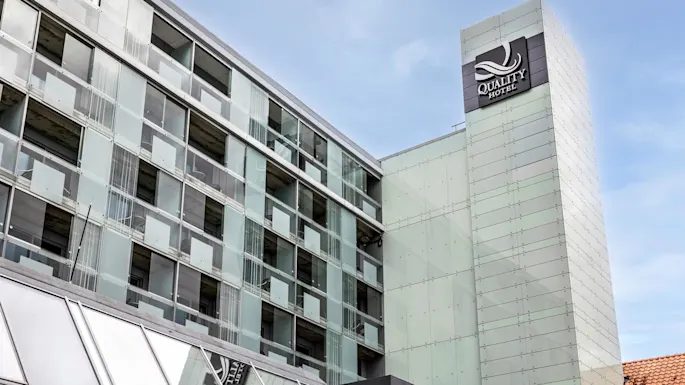 A modern glass building displaying the sign "QUALITY HOTEL" rises against a clear blue sky, emphasizing urban architecture with geometric lines and reflective panels.