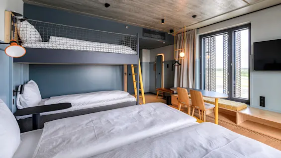 Et moderne hotelværelse med en køjeseng, enkeltseng og siddeområde ved store vinduer. Træelementer og blød belysning fremhæver det stilfulde, minimalistiske design.