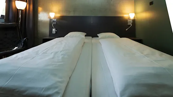 Two neatly made single beds with white linens are arranged side by side against a dark headboard, illuminated by wall-mounted lamps in a softly lit room with curtains and a chair.