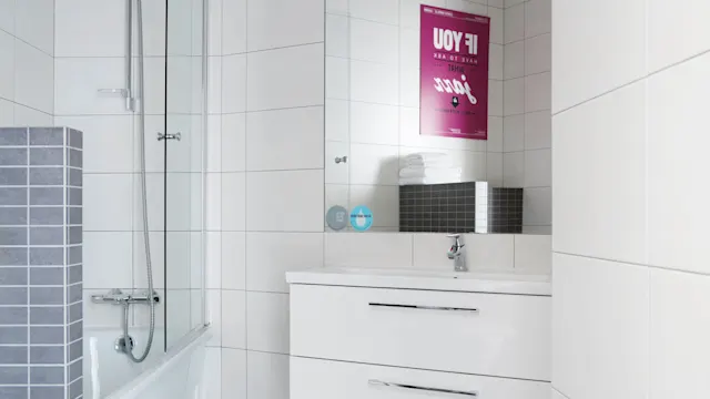 A modern bathroom has a white sink cabinet with a faucet beneath a rectangular mirror. A pink poster reads, "IF YOU HAVE TO ASK WHAT JAZZ IS, YOU'LL NEVER KNOW," reflecting on tiles.
