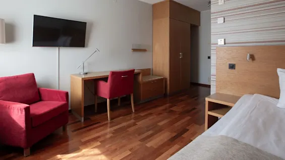 Red chair sits near a wooden desk with a lamp, opposite a wall-mounted TV. Adjacent is a bed with a wooden headboard, in a modern room with striped wallpaper.