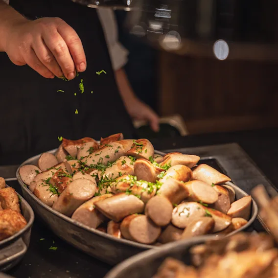 En koks hånd som strør ferske grønne urter over et varmt buffetfat fylt med julepølser.