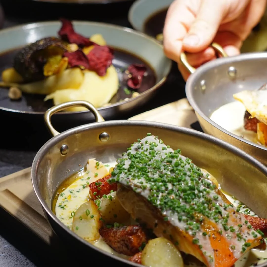 A metal dish holds salmon topped with herbs, resting on potatoes and sauce. A hand grasps the dish's handle, surrounded by other plated meals on a dark surface.