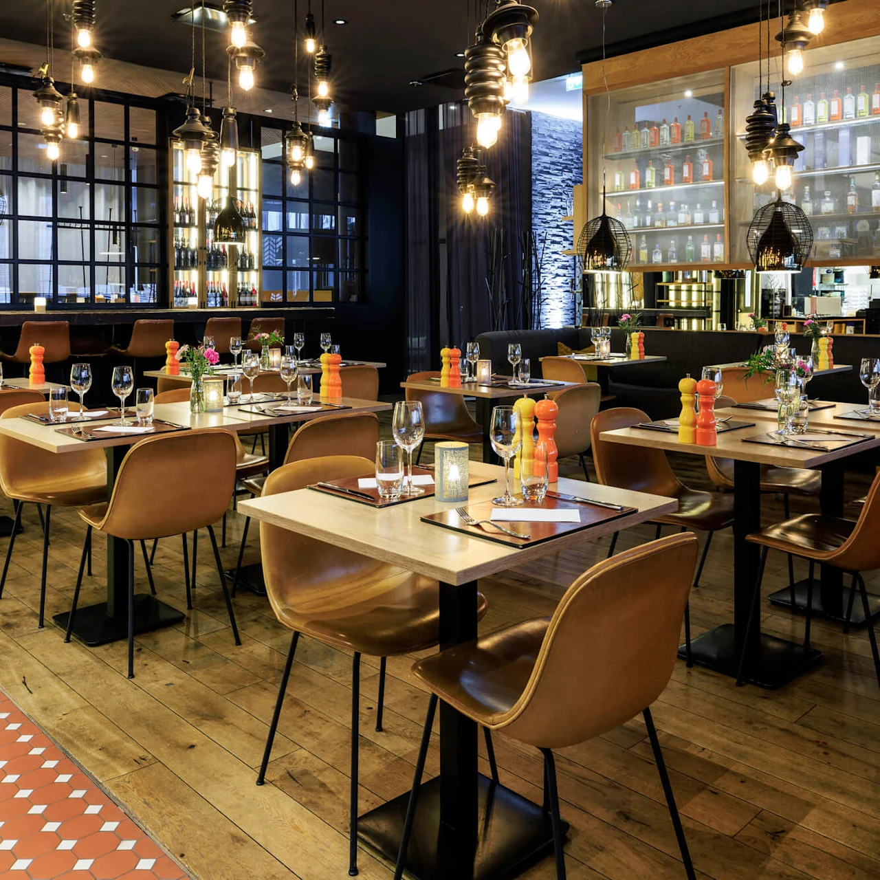 Tables are set with glassware and colorful shakers in an elegant restaurant. Warm lighting from hanging fixtures illuminates shelves lined with bottles, creating a cozy and inviting atmosphere.