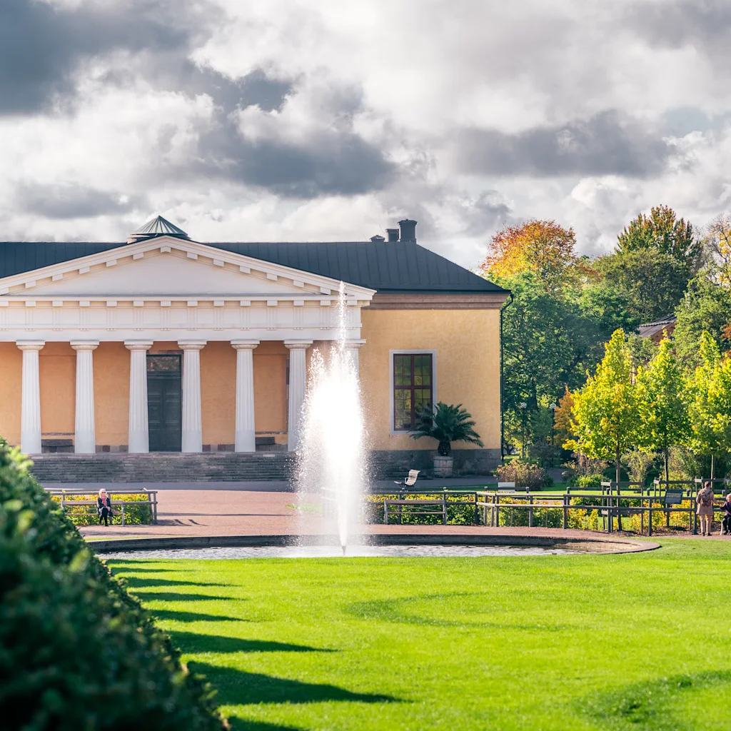 A grand building with a fountain in The Botanical Garden Uppsala, surrounded by vibrant autumn trees and people relaxing.
