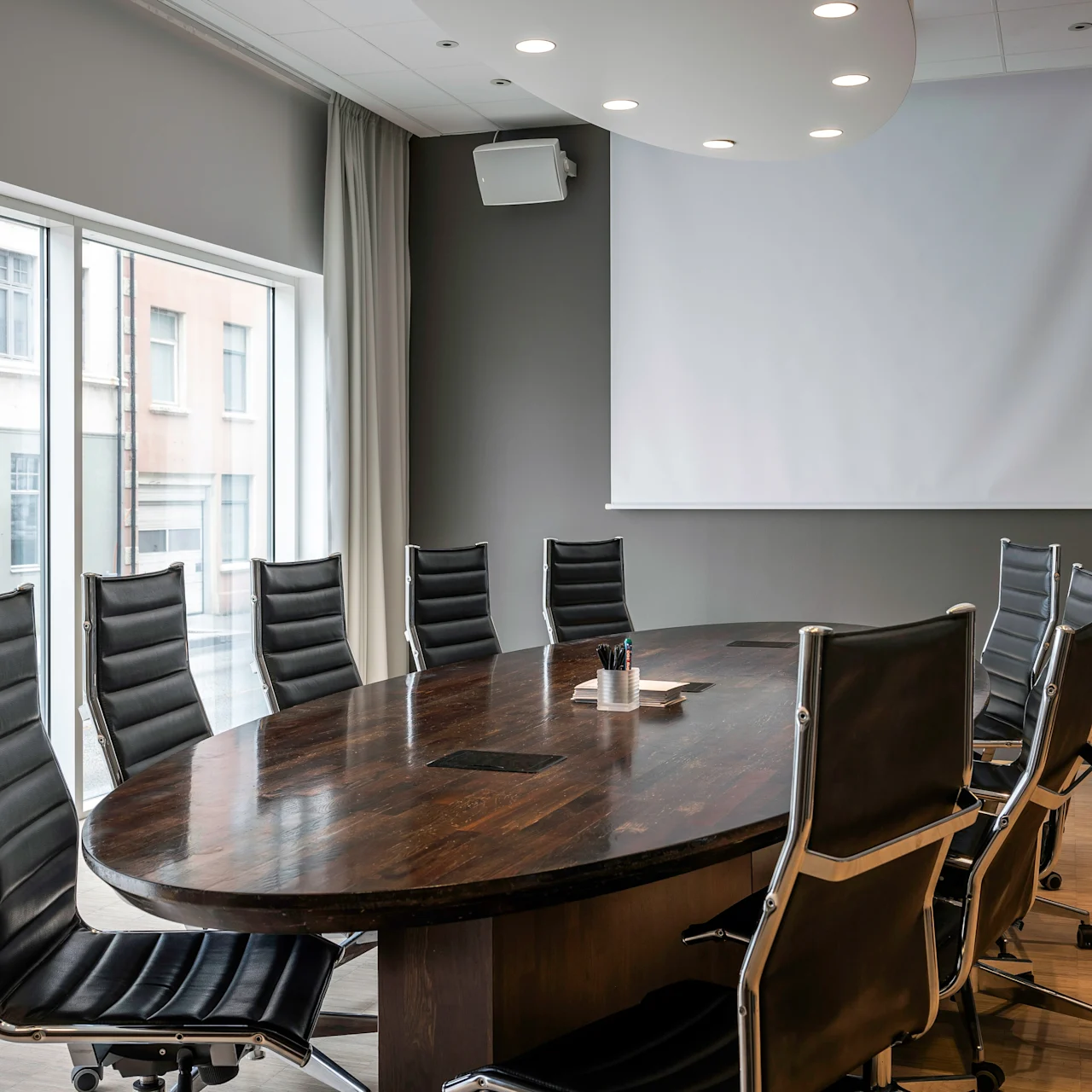 Ett stort konferensbord av trä omgivet av svarta stolar står i ett rum med en projiceringsskärm och ett stort fönster med utsikt över en gata och byggnader.