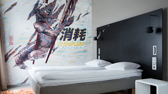 Bed with white linens, positioned against a black headboard with hanging lamps. Wall art depicts an armored figure with Japanese characters: "消耗されゆくもの". Room features an orange-patterned carpet.