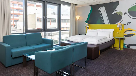 A hotel room with a teal sofa set faces a large window. A double bed is near a wall mural of a face and sunglasses.