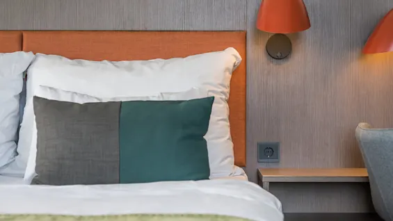 A bed is neatly made with white pillows and a gray-green decorative pillow, beside a wall with two modern red lamps and an electrical socket. 