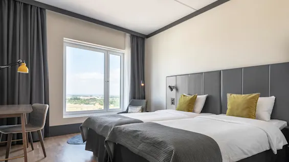 A neatly made bed with gray headboard and green pillows sits in a modern room. A large window overlooks a distant landscape. A desk and chair are by the window.