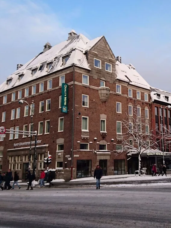 Ett snötäckt tegelhotell står vid ett trafikerat gatuhörn, omgivet av fotgängare och snötäckta träd. Skyltar lyder "Quality Hotel" och "Kurs & Konferansesenter".