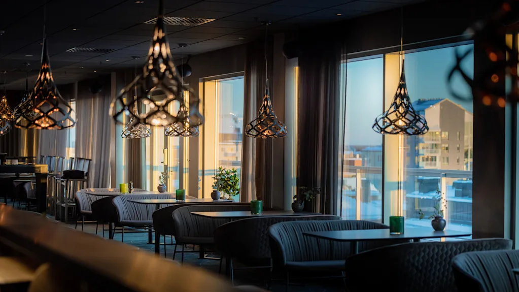 Stiliga hänglampor hänger ovanför bekväma sittplatser i en elegant, modern restauranginteriör, solljus strömmar genom stora fönster med utsikt över stadens byggnader.
