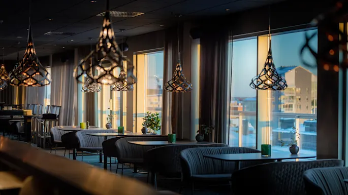 Stilige pendellamper henger over koselige sitteplasser i en elegant, moderne restaurantinteriør, sollys som strømmer gjennom store vinduer med utsikt over bybygningene.