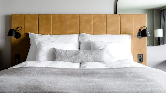 A neatly made bed with white pillows and a beige blanket rests against a tan upholstered headboard, flanked by black wall lamps in a modern room with a mirror.