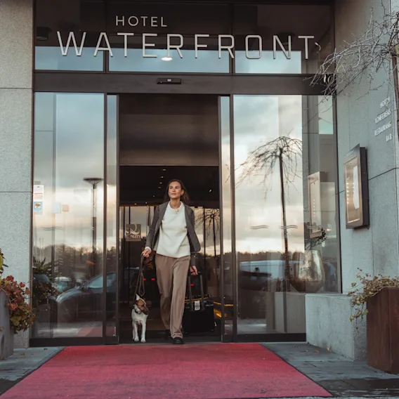 En person och deras hund lämnar ett hotell som heter Waterfront, går på en röd matta med bagage.
