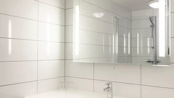 Et rent, moderne badeværelse med hvide flisebelagte vægge, en vask og et spejl med belyste sider, der reflekterer en bruser.